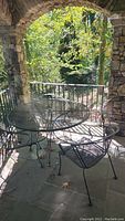 Black metal mesh round outdoor table with umbrella hole and four matching chairs on stone porch with stone pillars and railing