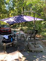 Full view of large levered umbrella opened with dark fabric, supported by a metal frame and single central pole. Located outdoors among patio furniture and trees.