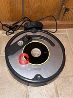 iRobot Roomba 630 vacuum robot with remote control and red brush cleaning tool on tiled floor near wooden wall.