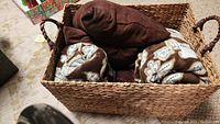 Photo showing large rectangular wicker basket filled with two brown pillows and three patterned throws.