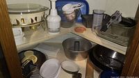 Photo showing a corner cabinet shelf with various kitchenware items including mixing bowls, angel food cake pan, muffin pans, glass baking pans, grater, and cookie press.