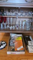 Wide view showing multiple shelves with varied glassware including stemware, steins, bowls, and cocktail glasses. Box in front contains bar tools and accessories, napkins visible in stack beside box.