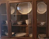 China cabinet displaying plates, saucers, bowls, cups, and serving tray with gold leaf design and trim.