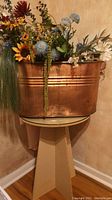 Copper tub filled with assorted faux flowers and greenery placed on a round particle board table with geometric base and glass top.