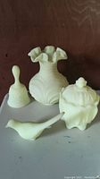 Four Fenton custard glass items on display: bell, fluted vase with ruffled top, lidded jar, and bird figurine.