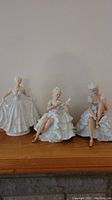 View of three white porcelain figurines on wooden surface, showing their full dresses and poses