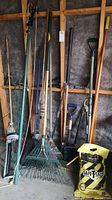 Photo showing various garden tools including rakes, shovels, broom, loppers, and bag of road salt against a wooden wall.