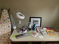 Wide view of sewing and quilting supplies on table including iron, books, pins, ruler, lamp, and framed print.