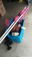 Blue plastic storage bin filled with various sporting items including basketballs, a badminton net with pink poles, golf balls, and an air pump. Two black dumbbells are placed outside the bin on the floor.