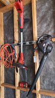 Photo showing the Black & Decker cordless string trimmer mounted on a wooden frame with an 18V battery charger and red battery pack alongside a long orange extension cord. The string trimmer is red and black with telescopic shaft and automatic spool feed cutting head.