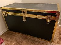 Front view of large rectangular textured black trunk with brass edges, brass latches on front, and lock in center.