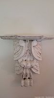 Front view of composite carved wall shelf mounted on wall, showing leaf and scroll carved details and distressed white paint finish