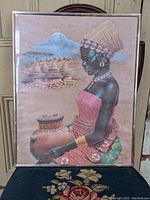 Full view of the framed print depicting the African tribal woman sitting with traditional attire, pot, and background elements, mounted in a silver metal frame.