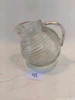 Angled side view of clear depression glass ball pitcher with horizontal ribbed Manhattan pattern, showing handle and spouted mouth.