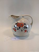 Front left angle view of the white ceramic ball pitcher with red poppy flower design and handle.
