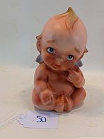 Front view of ceramic Kewpie doll still bank, showing seated baby figure with a finger near mouth and distinct hair tuft.