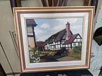 Frontal photo of framed oil painting depicting Tudor style house outdoors, showing white and dark timber wall sections, thatched roof, chimney and garden with flowers.