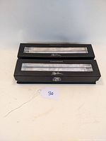 Two black boxes with clear plastic windows showing the candlestick holders inside. Boxes are branded with 'Oleg Cassini' and labeled 'Crystal Candlestick'.