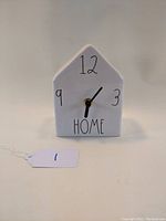 Front view of white ceramic house-shaped clock showing black numerals 12, 3, 9 and the word 'HOME' with black hands.