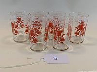 Six clear glasses with a red tulip design and gold trim, arranged with some staggered in front and others behind showing full set and detail.