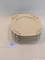 Stack of 8 ivory square plates with decorative Riviera pattern, visible border design, one plate chipped at corner