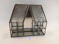 View of the front and one side of the vintage house-shaped curio display with glass shelves and mirrored back, showing the textured roof glass panels and metal frame.