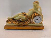Front view of vintage Taranis mantle clock showing reclining female figure with gold painted drapery and clock face inset on right side.