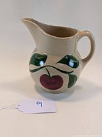 Front and side view of the ceramic pitcher showing the apple pattern design with red apples and green leaves on beige background.