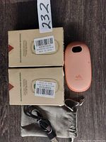 Two rechargeable peach/coral hand warmers, their original cardboard boxes, black USB cable and grey cloth pouch laid out on a dark wood surface.