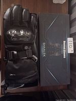 Photo showing one black heated glove with carbon fiber knuckle protection and adjustable strap alongside its original Shaalek branded box.