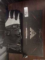 Photo showing one pair of black heated gloves with reflective panels and control button, placed on a black box labeled as Bacialeri Heated Gloves.