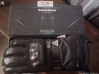 Photo of large black Shaalek heated gloves with knuckle protection and control buttons shown alongside the product box.