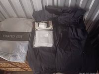 Black women's heated vest laid out flat alongside its packaging and a small zipped pouch containing accessories.