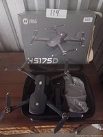 Photo showing Holy Stone HS175D drone with folded arms inside a black carrying case, original box placed behind. Accessories including a propeller guard and other items in plastic are visible.