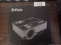 Black Jinhoo projector in original box with brand and projector illustration on the lid, marked with lot number 102.