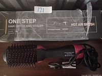 Photo of a One Step Hair Dryer and Styler Hot Air Brush with bristles on brush head, black and pink handle, and power cord attached lying on a seating surface, box visible in background with product information.
