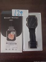 Photo showing black sport smart watch in box with silicone strap and physical buttons, with label '129' present