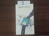 Photo of the boxed smart watch showing the front of the retail packaging with a white background and image of the watch with black strap.