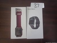 Photo of smart watch with purple silicone band placed inside original white box packaging. The box lid shows an image of the watch with a black band and the text 'Smart Watch'.