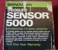 Photo of the original box labeling the item as a Brinkmann Treasure Sensor 5000 Discriminating T/R Metal Detector with warranty information and features.