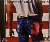 Close-up photo of Bruce Springsteen's Born in the USA CD cover showing a man from the back wearing jeans with a red cap in pocket against an American flag background.