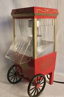 Front view of red and gold decorative popcorn maker with clear acrylic chamber and front chute, showing wheels and internal popping mechanism.