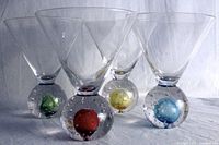 Four clear art glass cocktail glasses with colorful bubble bases arranged in a row showing red, green, yellow, and blue globes encased in heavy clear handblown glass bases with bubbles.