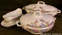 Three-piece set showing oval serving dish with lid, round serving dish with lid behind, and sauce boat with attached underplate to the left. All have colorful floral patterns and gold trim.