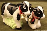 Pair of black and white ceramic cow-shaped salt and pepper shakers with red ribbons and gold bells around their necks, showing front and side views.