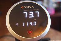 Close-up of the Anova sous vide cooker digital display showing current temperature 73.7°F, set temperature 114.0°F, with Bluetooth icon.
