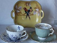 Wide shot of the Tillowitz yellow serving plate with parrots, Coalport blue and white bird teacup and saucer, and Aynsley light green and gold trim teacup and saucer together.