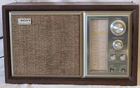 Full front view of the vintage Sony AM/FM radio showing wooden veneered cabinet, fabric speaker grille with Sony badge, and control panel with tuning dial and knobs.