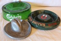 Lot showing all three vintage ashtrays: pewter cowboy hat, green glass Carlsberg beer, and ceramic John Smith's Bitter by Wade.