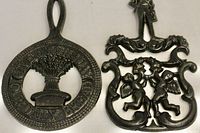 Top view of both vintage cast iron trivets side by side: left trivet circular with wheat sheaf and inscription; right trivet ornate with two cherubs.